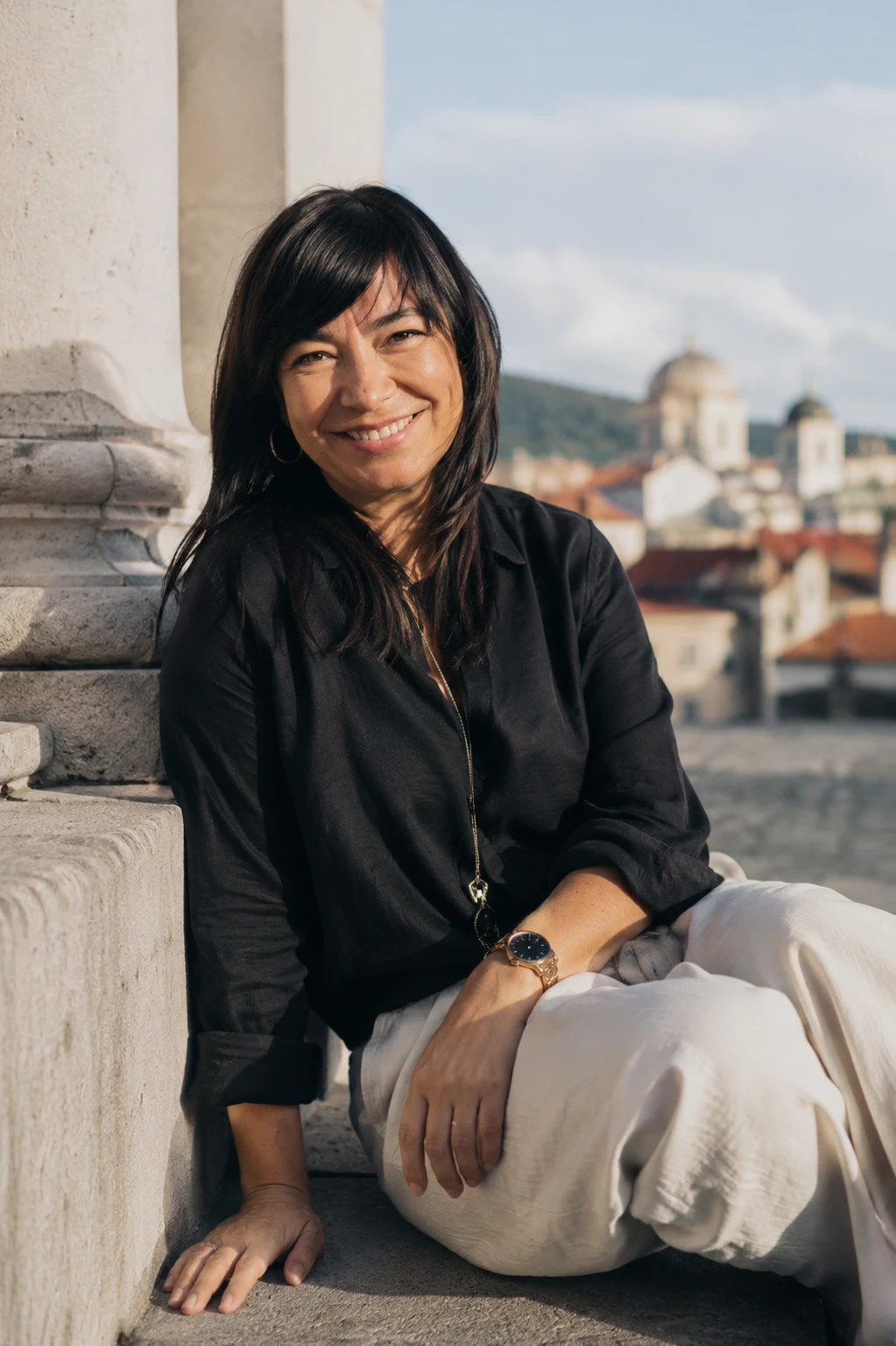 Nataša Popović smiling, seated against a stone column with an old town skyline behind her